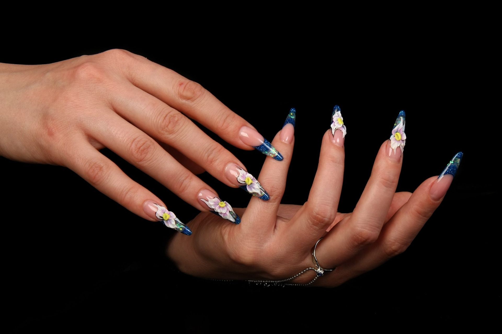 Manos con uñas largas y pintadas. Manicura francesa con puntas azules y diseños florales blancos. Fondo negro.