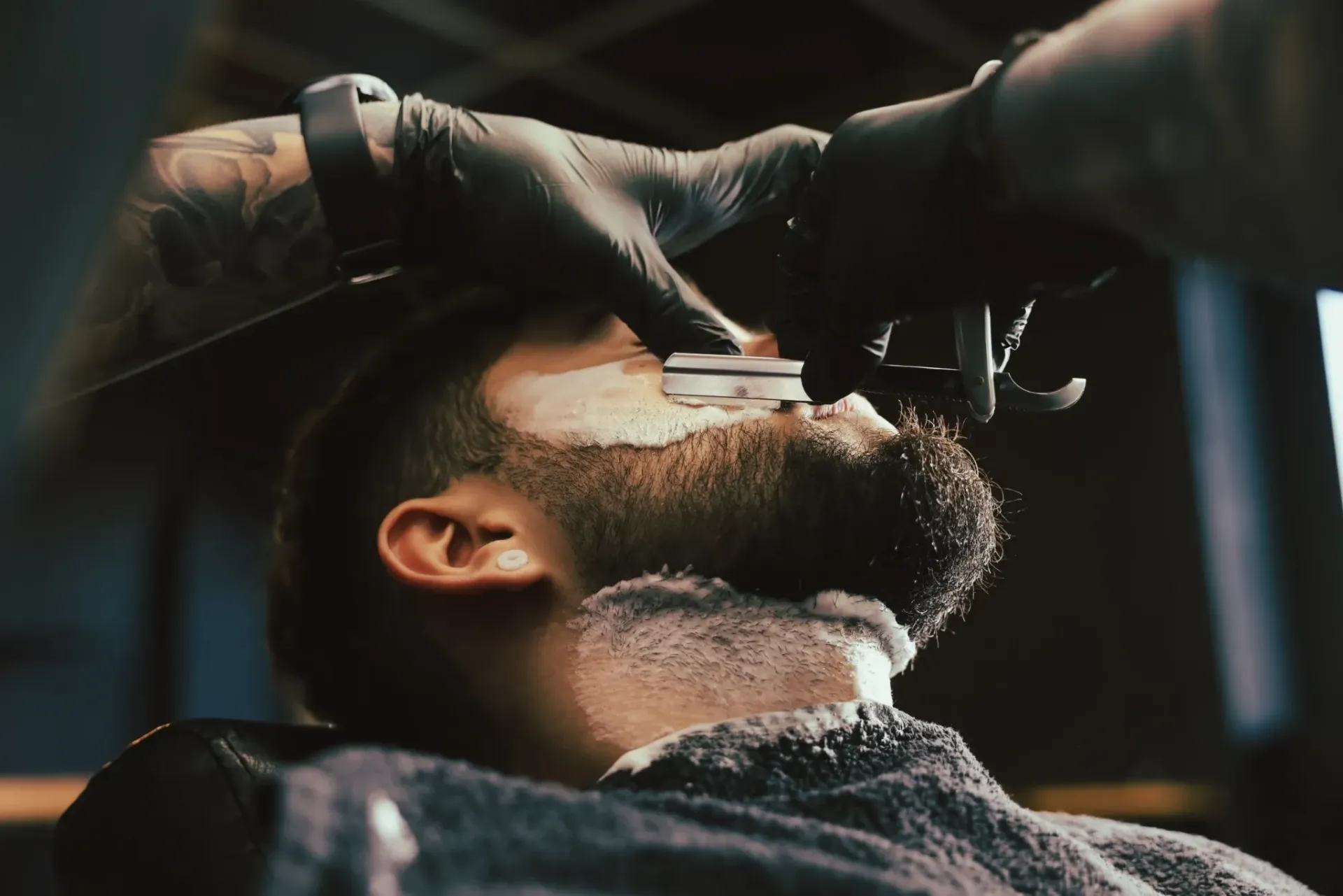 Un barbero afeita la barba de un cliente con una navaja. El cliente está en una barbería, con guantes negros.