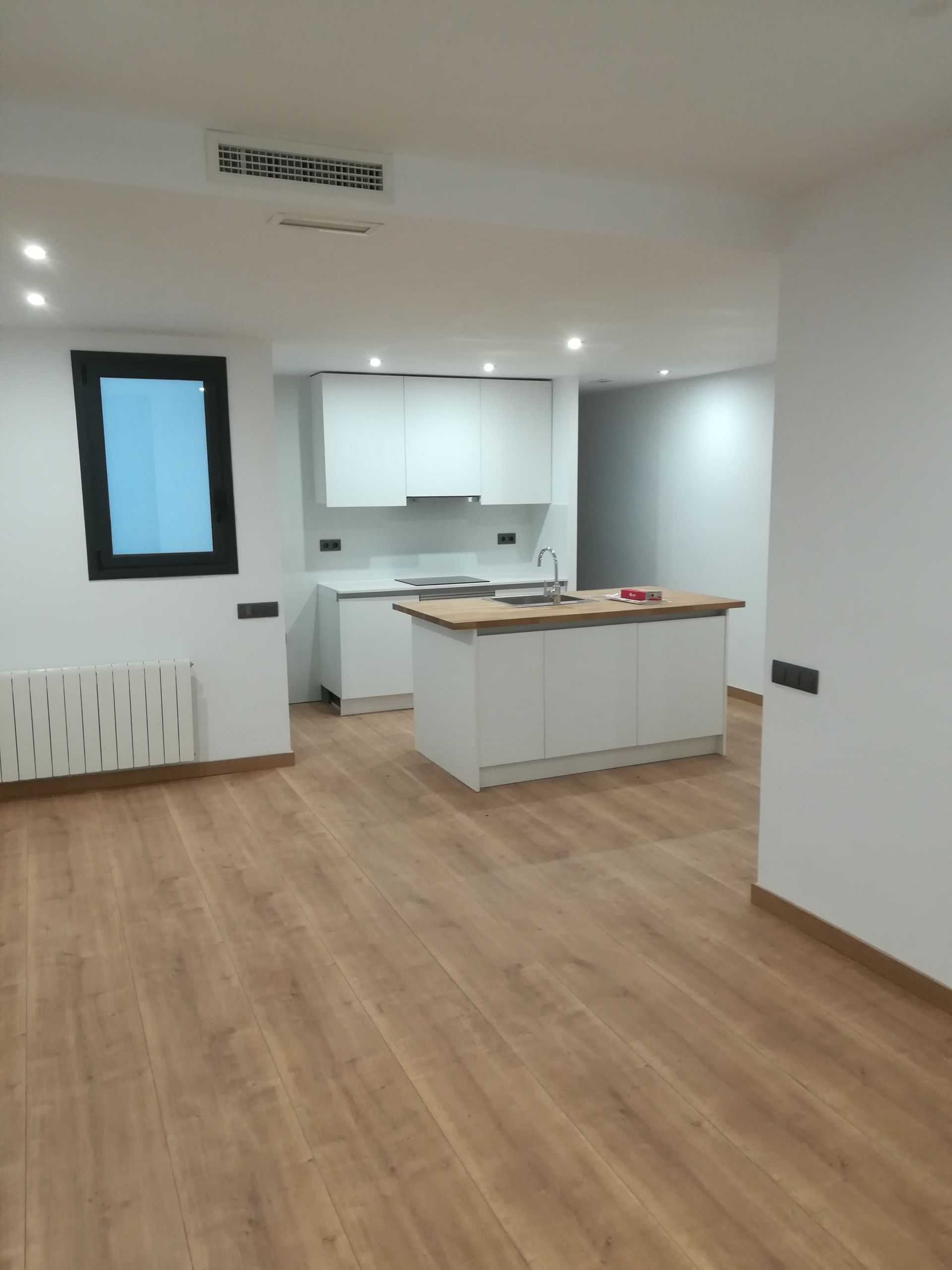 Cocina moderna vacía con armarios blancos, isla, suelo de madera y una ventana pequeña.