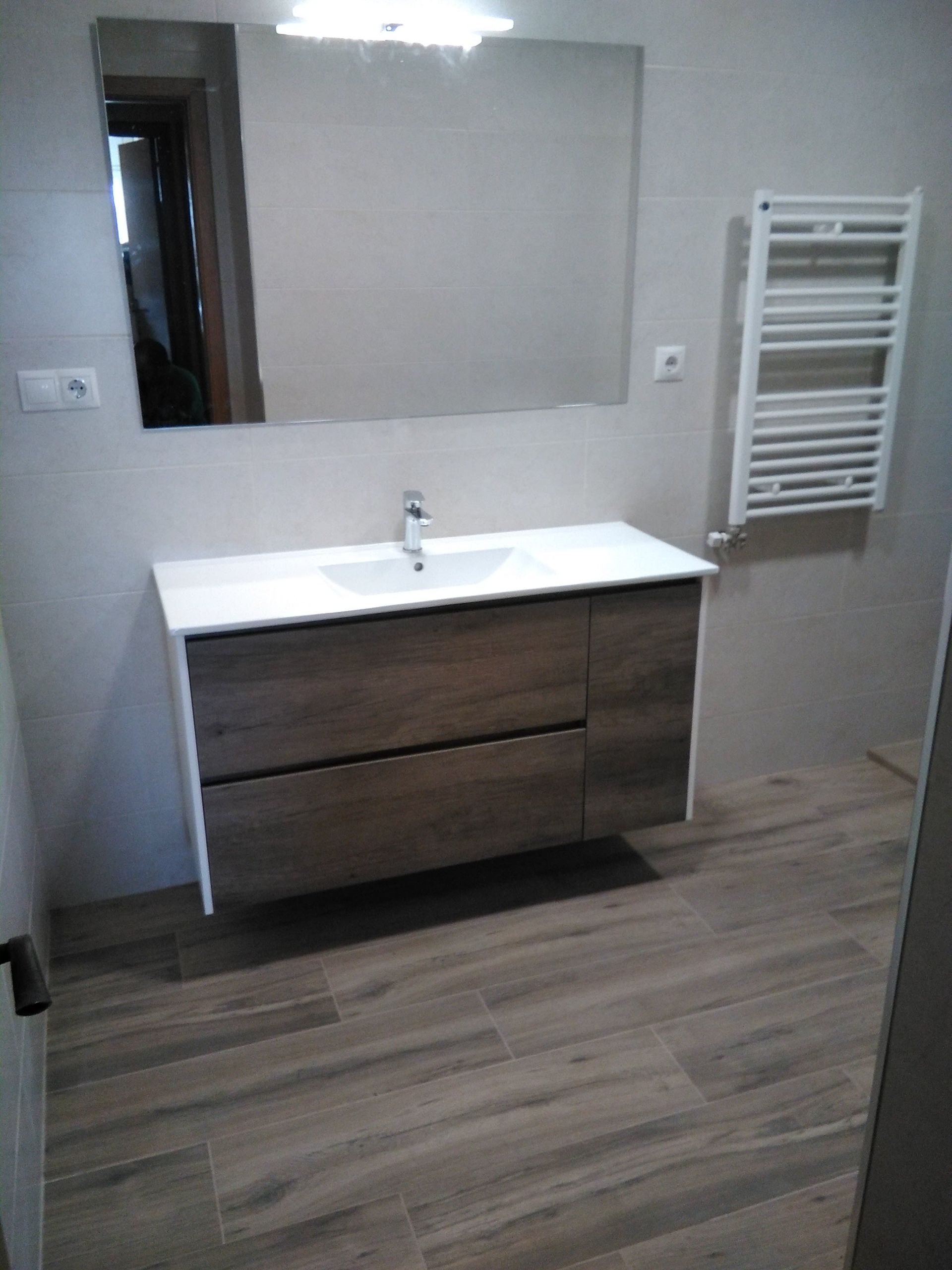 Mueble de baño moderno con espejo, lavabo, mueble de madera y toallero calefactable de pared, sobre paredes grises.