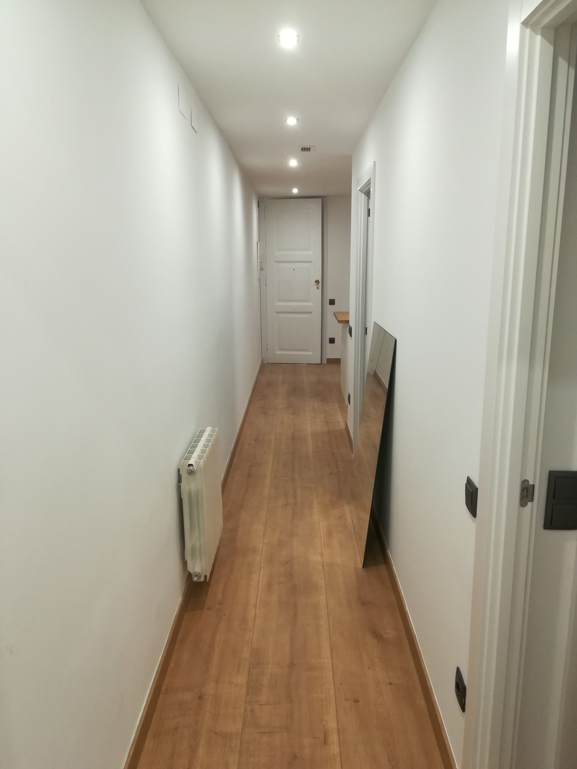 Pasillo estrecho y blanco con suelo de madera, radiadores, luces en el techo y una puerta al fondo.