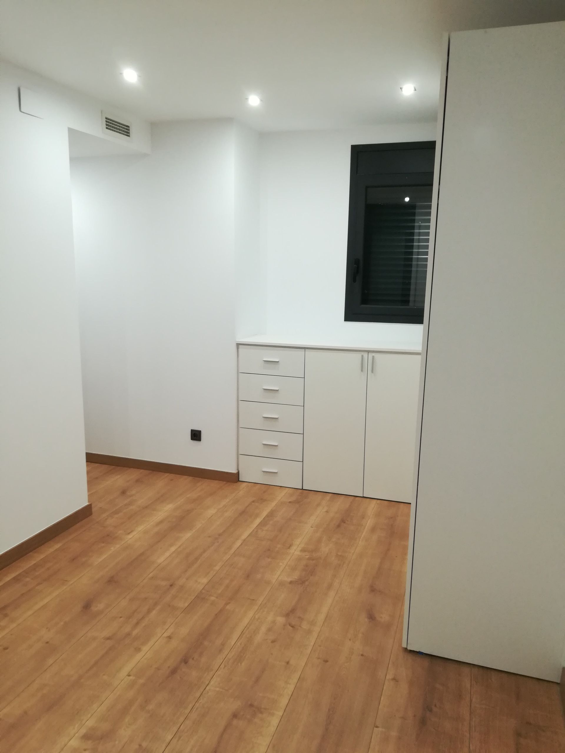 Habitación blanca vacía con suelo de madera, cajones empotrados y una ventana oscura