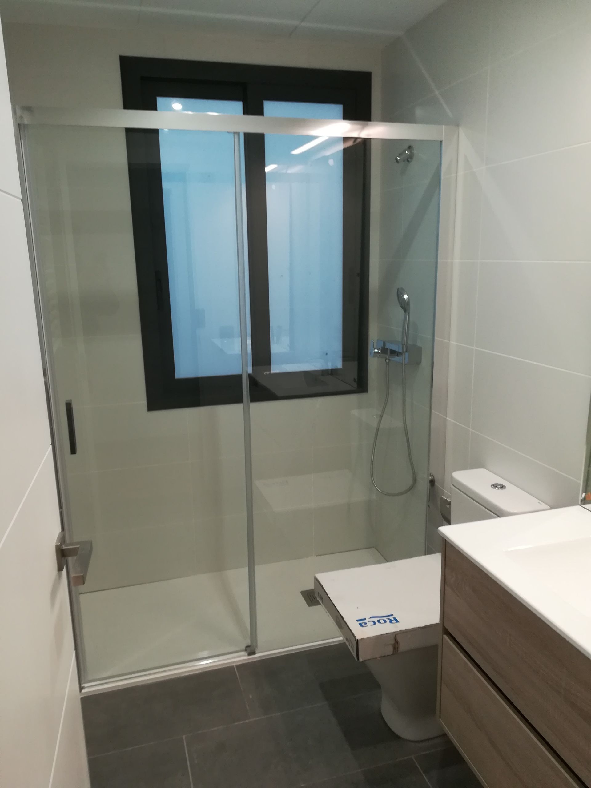 Baño moderno con mampara de ducha de cristal, inodoro, ventana y mueble lavabo