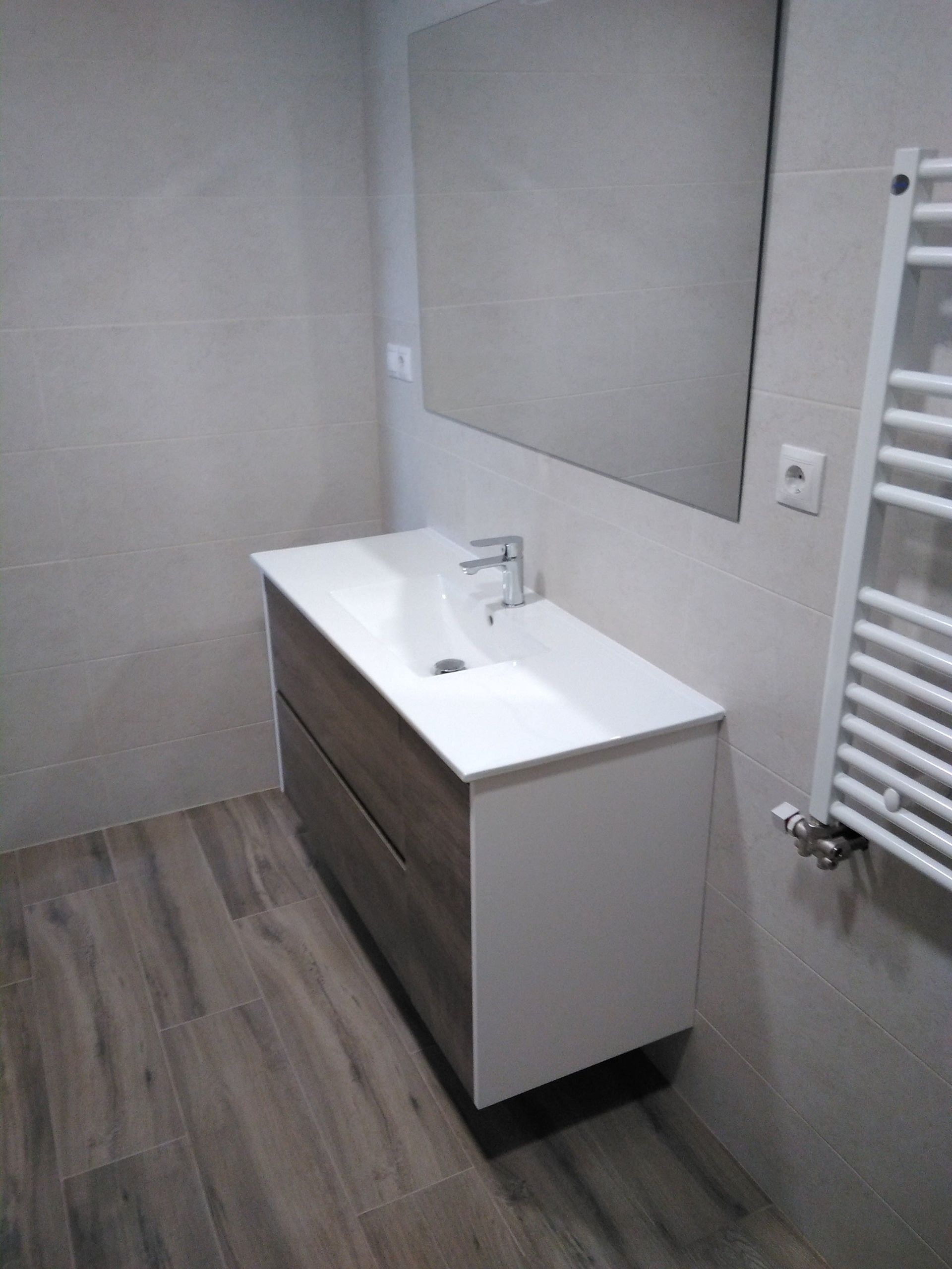 Mueble de baño moderno con lavabo blanco, armario de madera, espejo y radiador toallero en paredes grises.