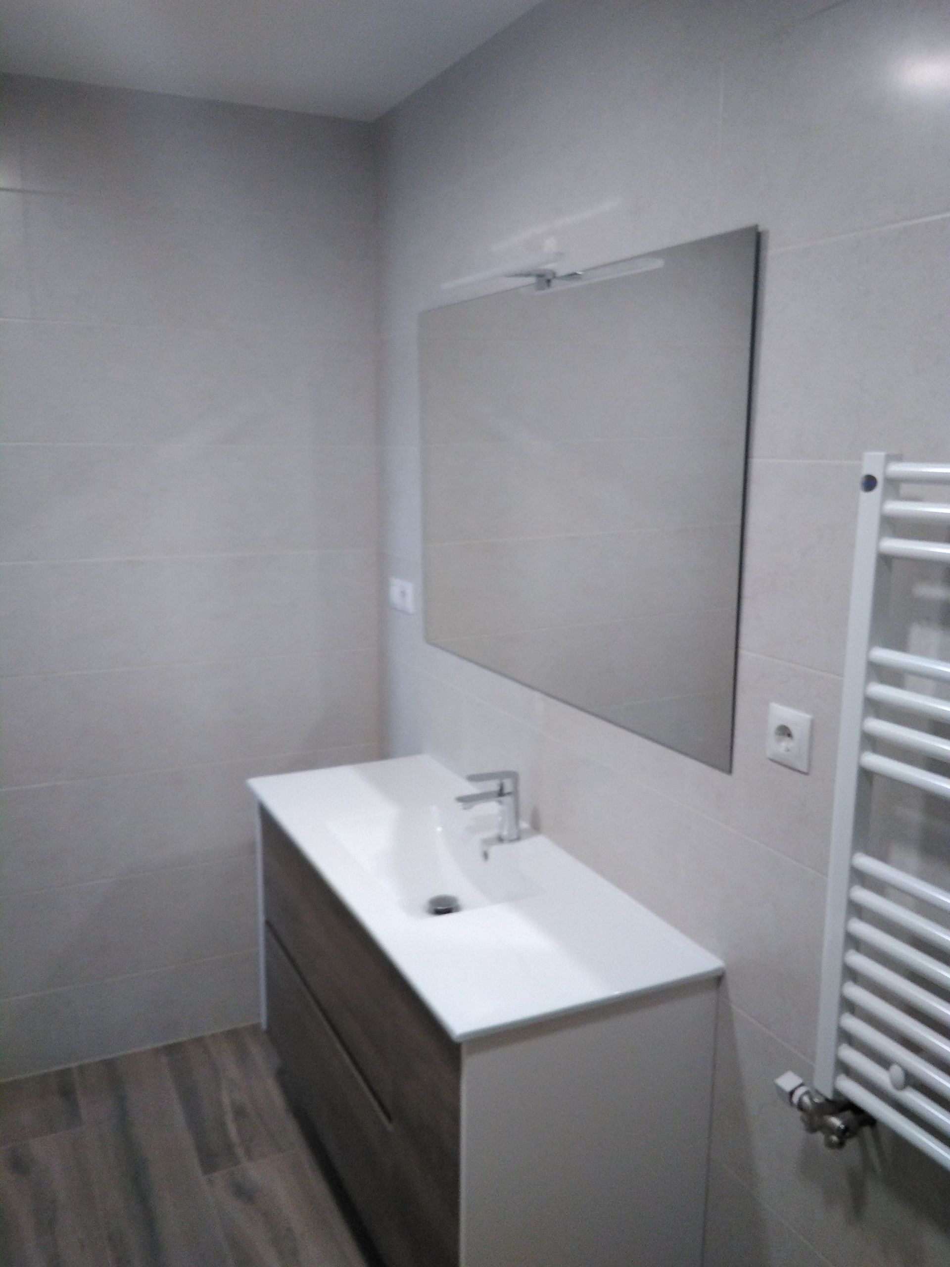 Baño moderno con lavabo blanco, mueble de baño oscuro, espejo grande y radiador toallero en paredes grises.