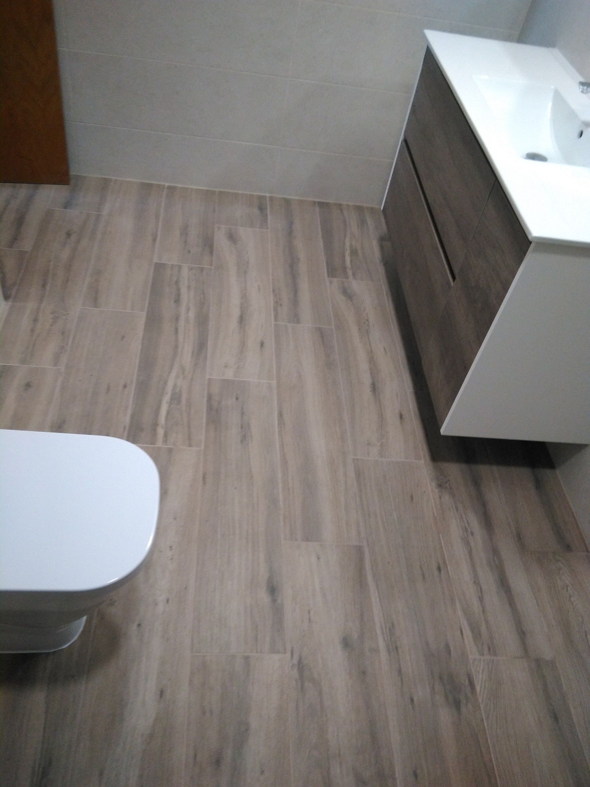 Baño pequeño con suelo de baldosas efecto madera, mueble de lavabo blanco y el asiento del inodoro a la vista en una esquina.