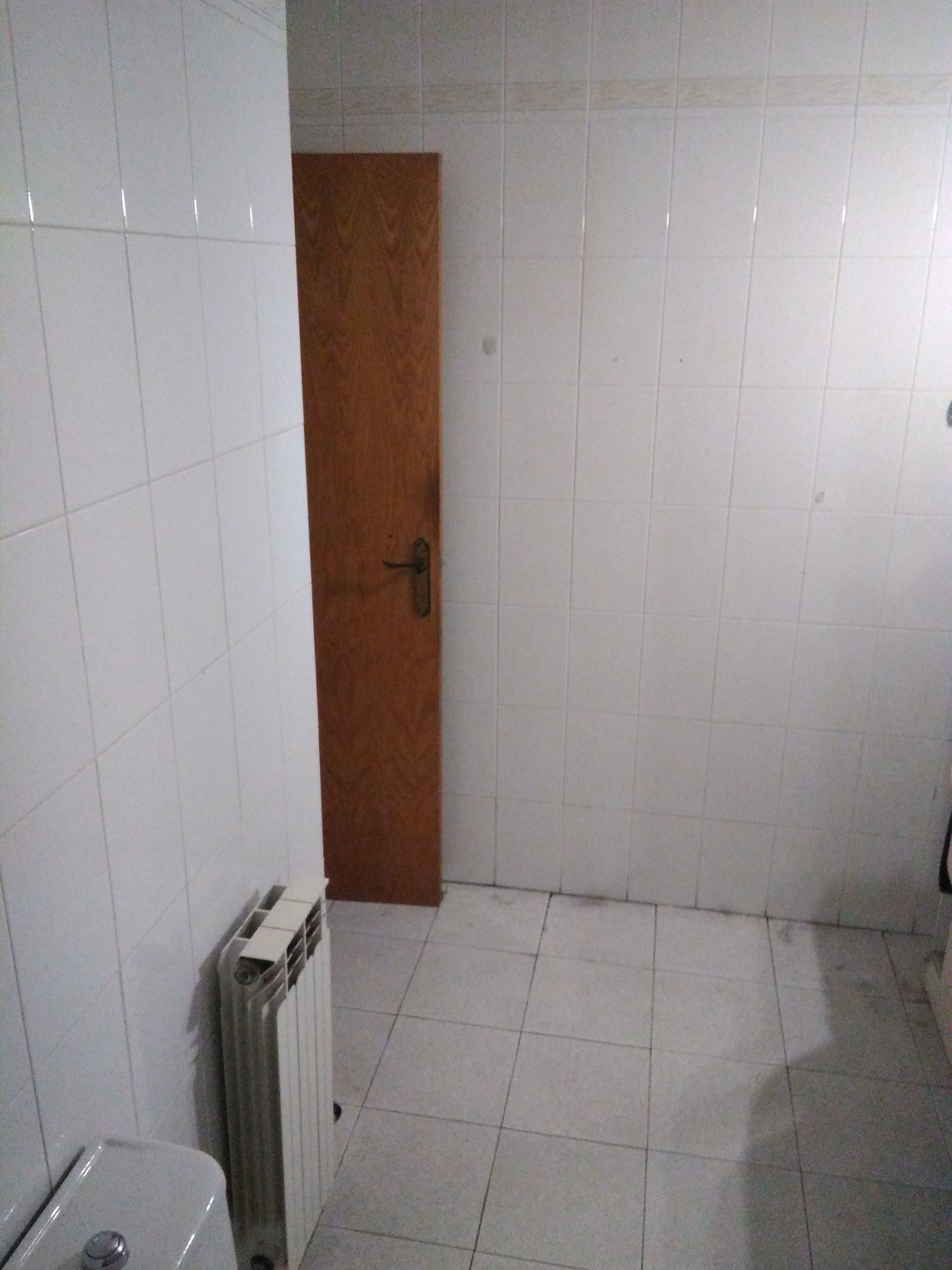Baño pequeño con azulejos blancos, una puerta de madera y un radiador de pared.