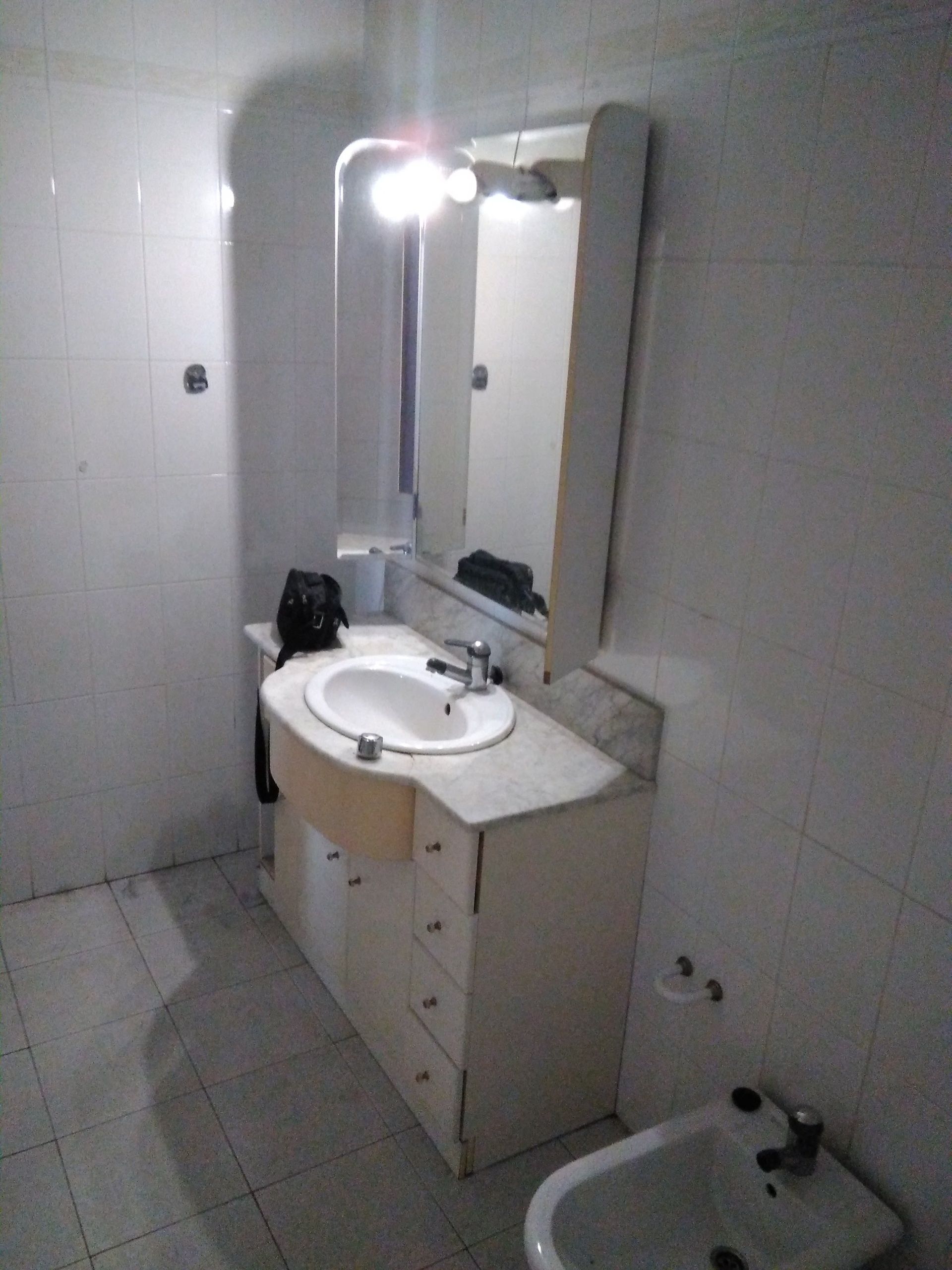 Baño pequeño alicatado, con lavabo, espejo e inodoro en una esquina; parece inacabado o desordenado.