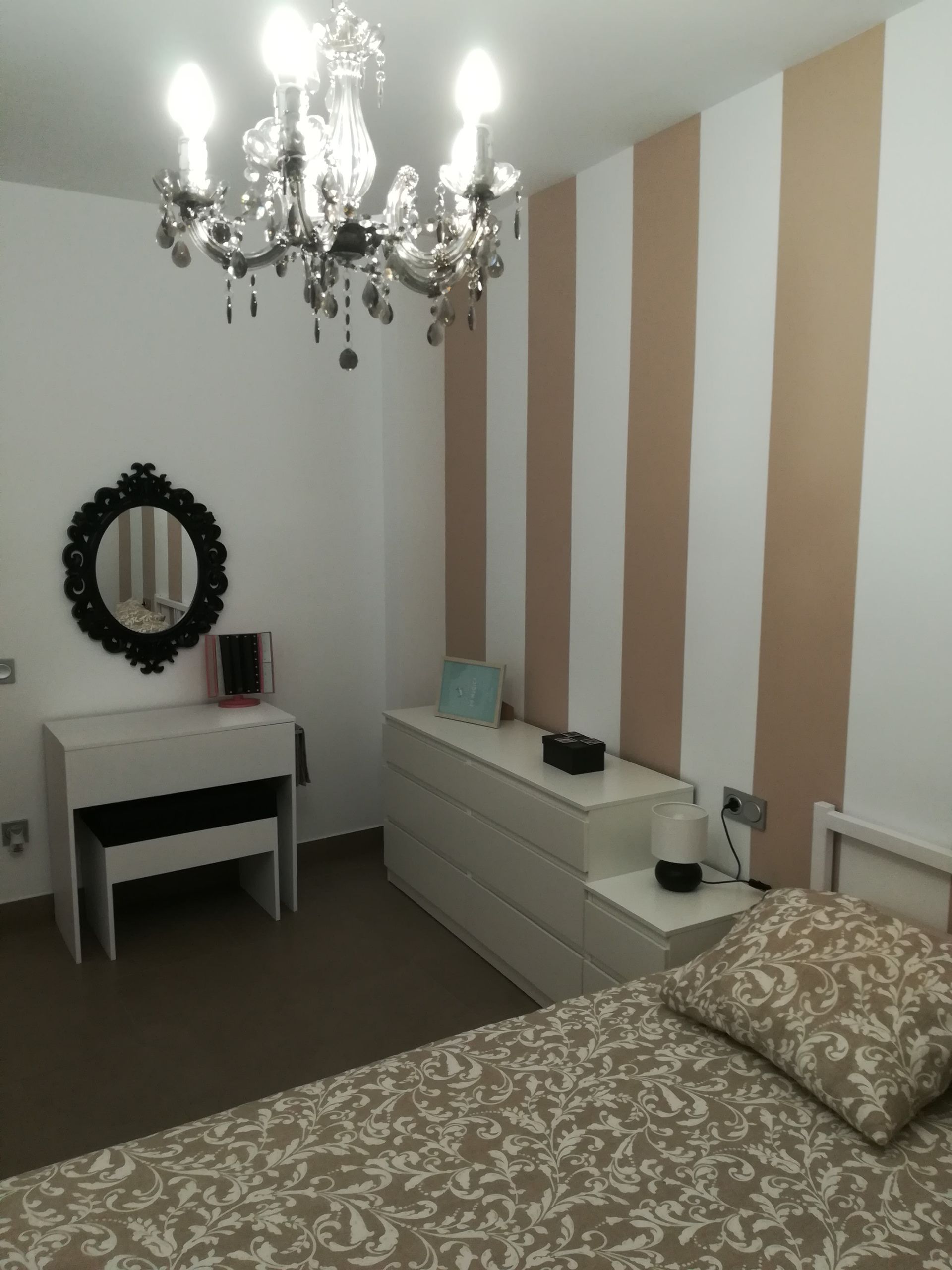 Dormitorio con lámpara de araña, pared decorativa a rayas, tocador, cómoda, espejo y cama