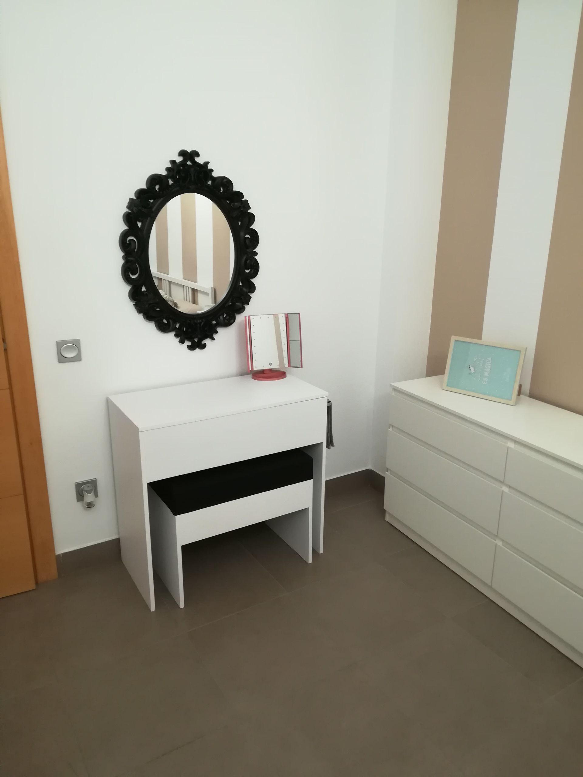 Dormitorio pequeño con un tocador blanco, un espejo ovalado negro, un banco y una cómoda junto a una pared color crema.