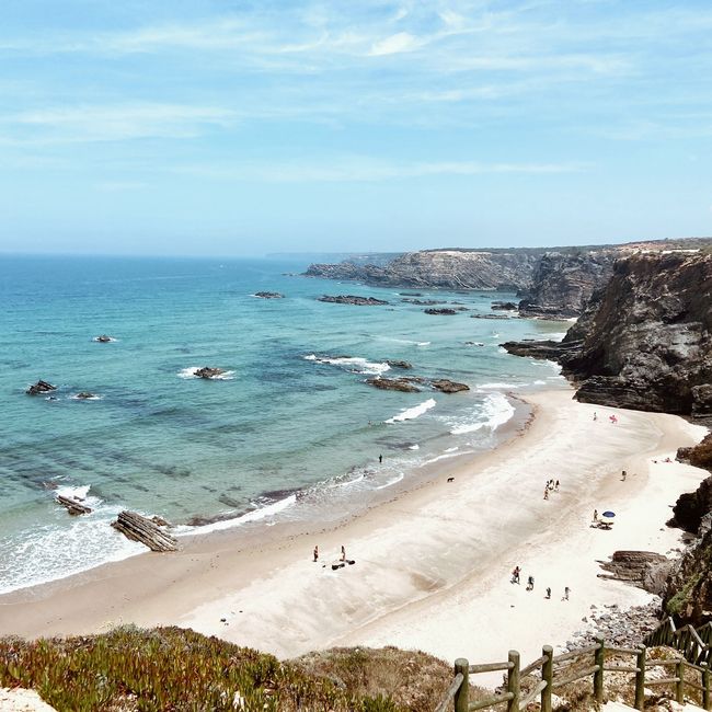 Enseada ensolarada com água turquesa, praia de areia, falésias rochosas e algumas pessoas caminhando pela costa.