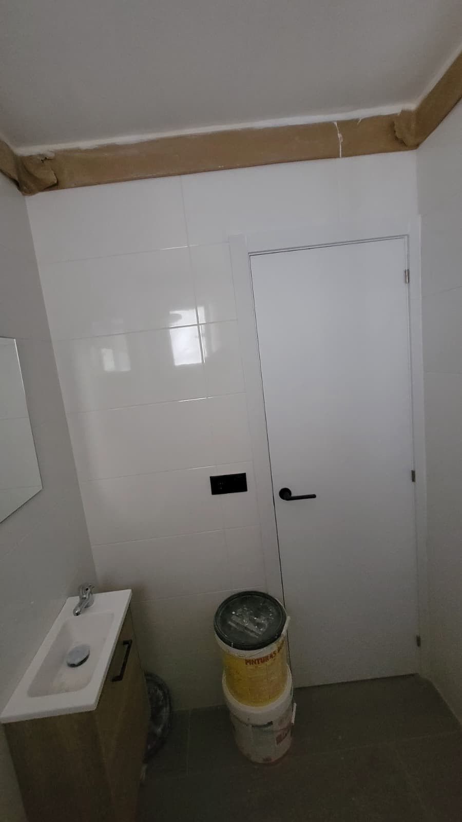 Un baño blanco con un pequeño tocador, una puerta blanca cerrada y una pila de dos cubos de pintura en el suelo gris.