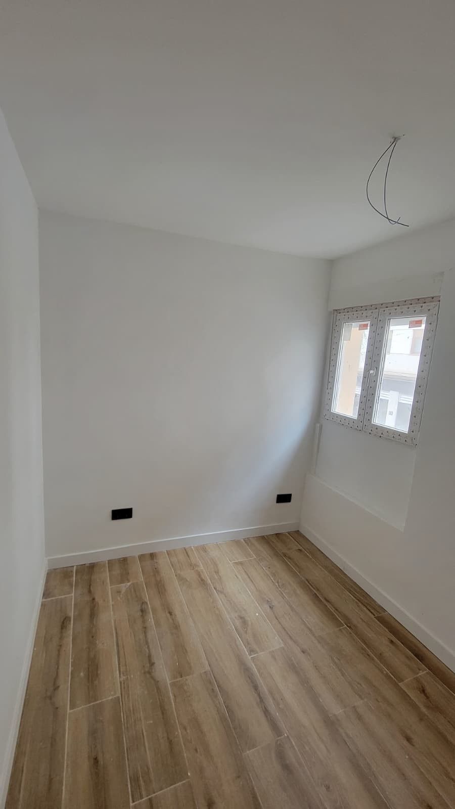 Una habitación pequeña y vacía con paredes blancas, suelo de baldosas que imitan la madera, dos enchufes y una sola ventana.