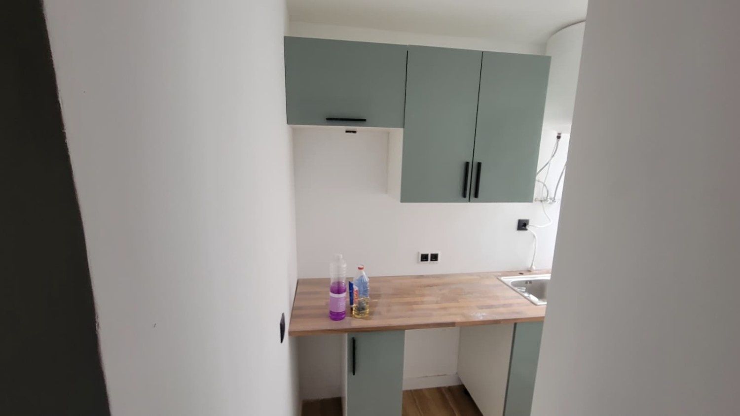 Una cocina pequeña y moderna con armarios de color verde salvia, encimeras de madera clara y paredes blancas.