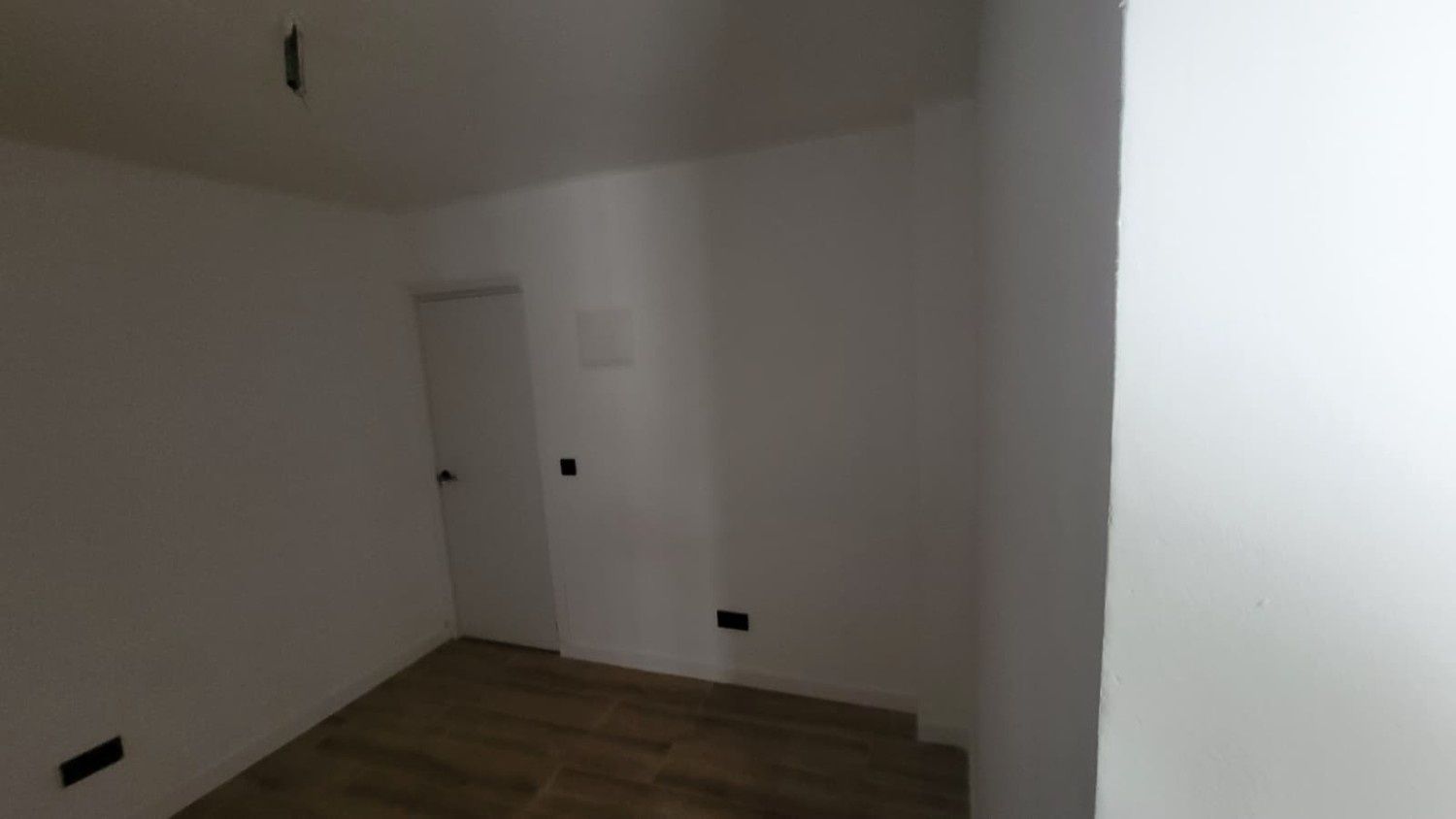 Una habitación vacía con paredes blancas, suelo que imita la madera, una puerta y enchufes en la pared.
