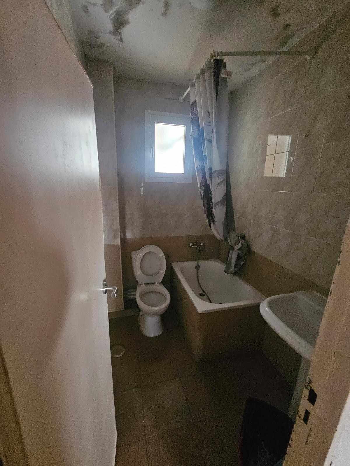 Un baño pequeño y anticuado con inodoro, bañera con cortina de ducha estampada y lavabo sobre suelo de baldosas marrones.