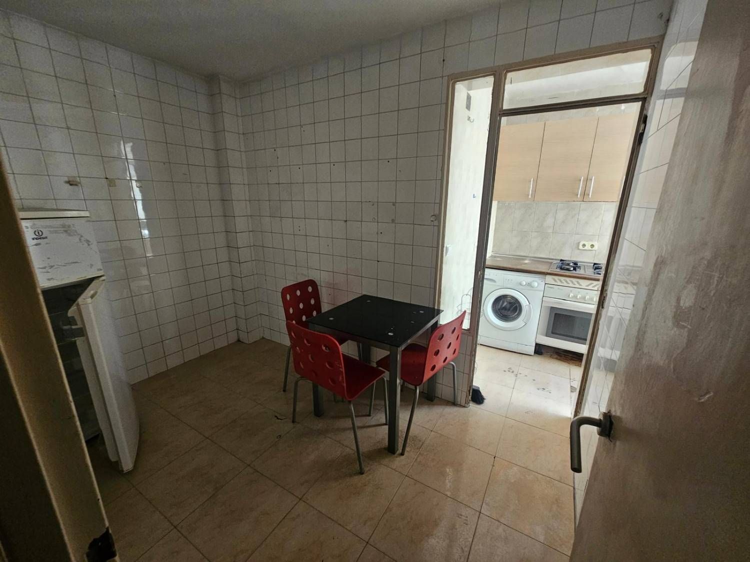 Una cocina con paredes alicatadas, una mesita con tres sillas rojas y una puerta 