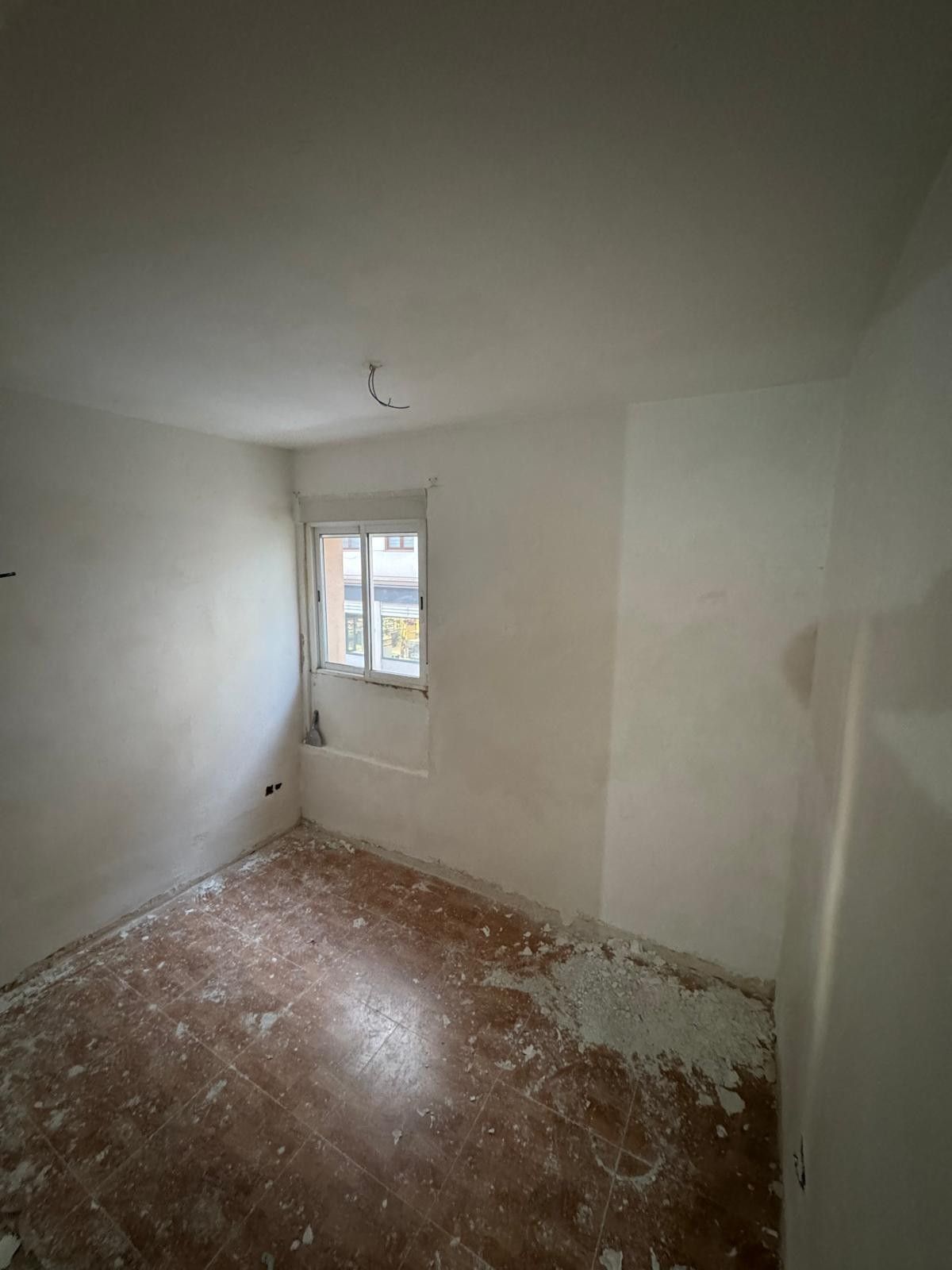 Una habitación pequeña y vacía en obras, con paredes blancas, una sola ventana y un suelo de baldosas cubierto de escombros.