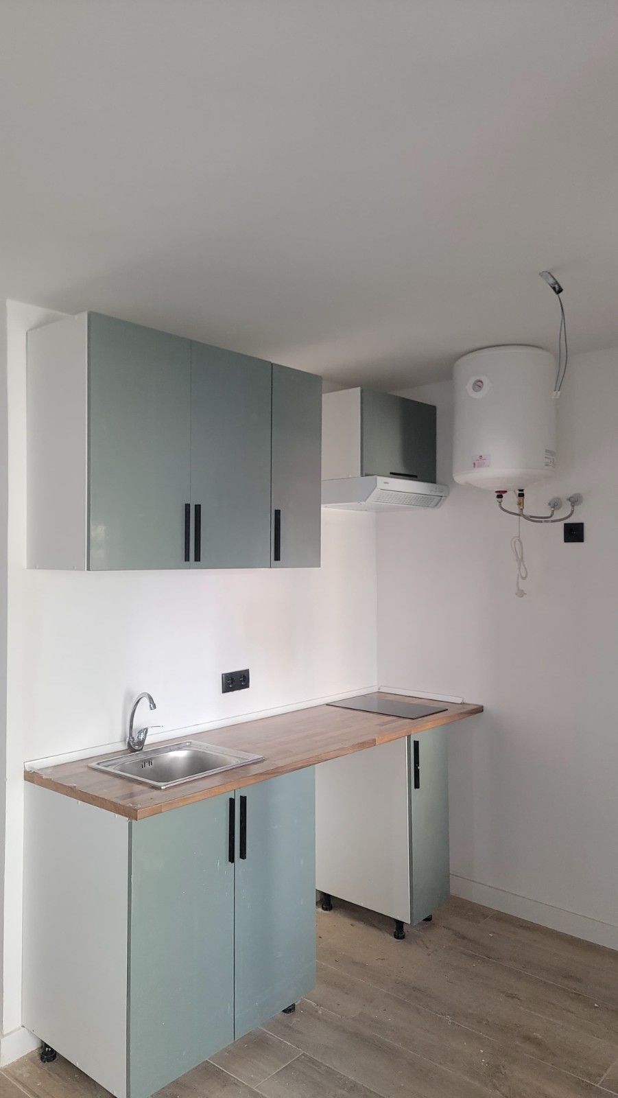 Una pequeña cocina con armarios azul claro, encimera de madera, fregadero de acero inoxidable y calentador de agua mural.