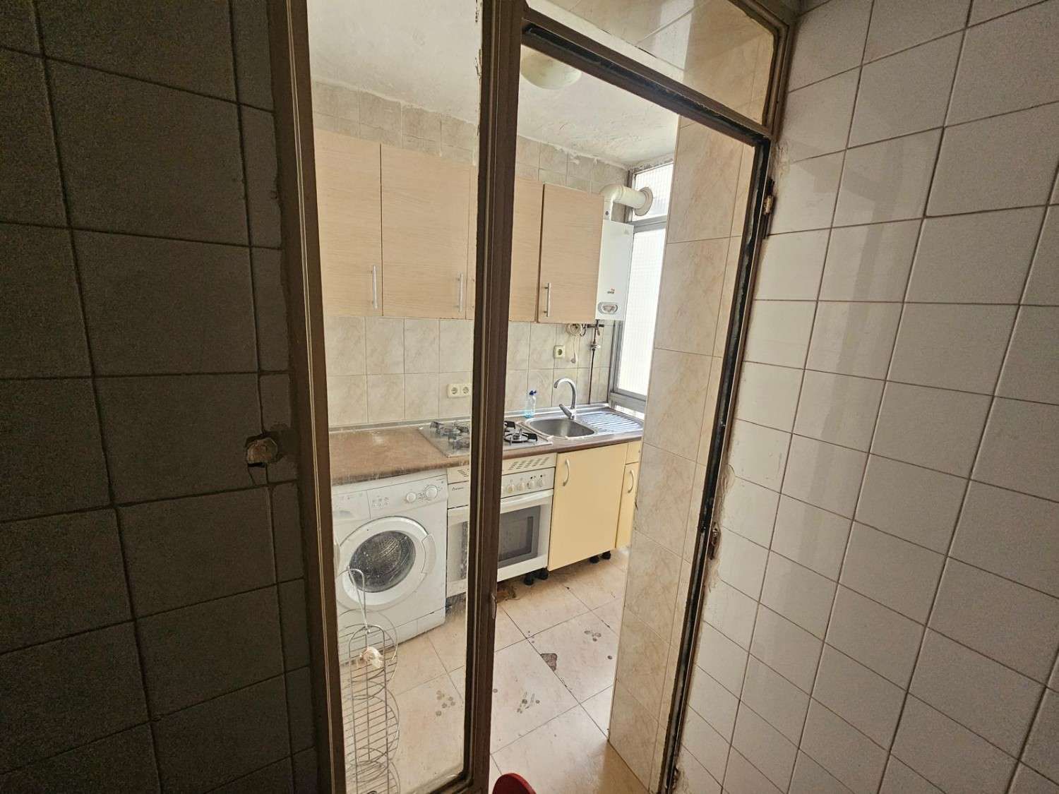 Una cocina vista a través de una puerta, con armarios de color claro, una lavadora, una estufa y paredes alicatadas.