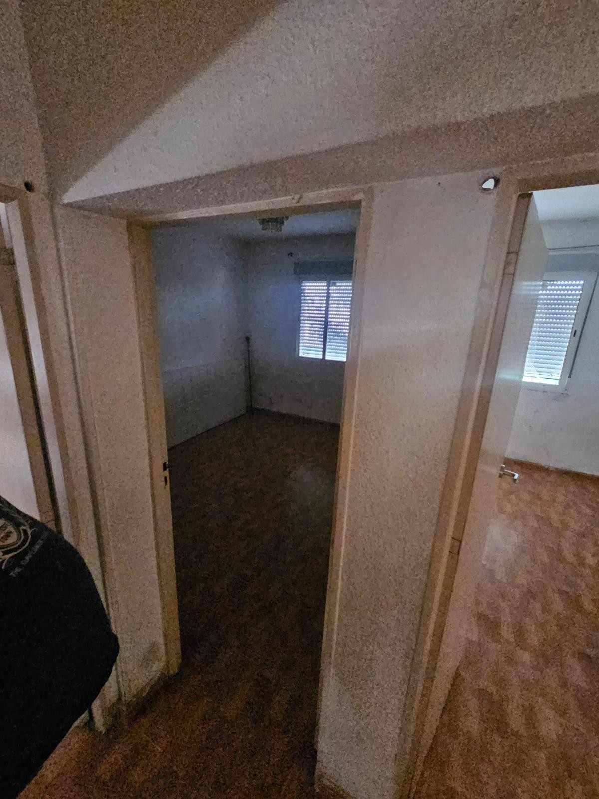 Vista a través de una puerta hacia una habitación vacía con suelo de madera y una ventana con persianas horizontales.