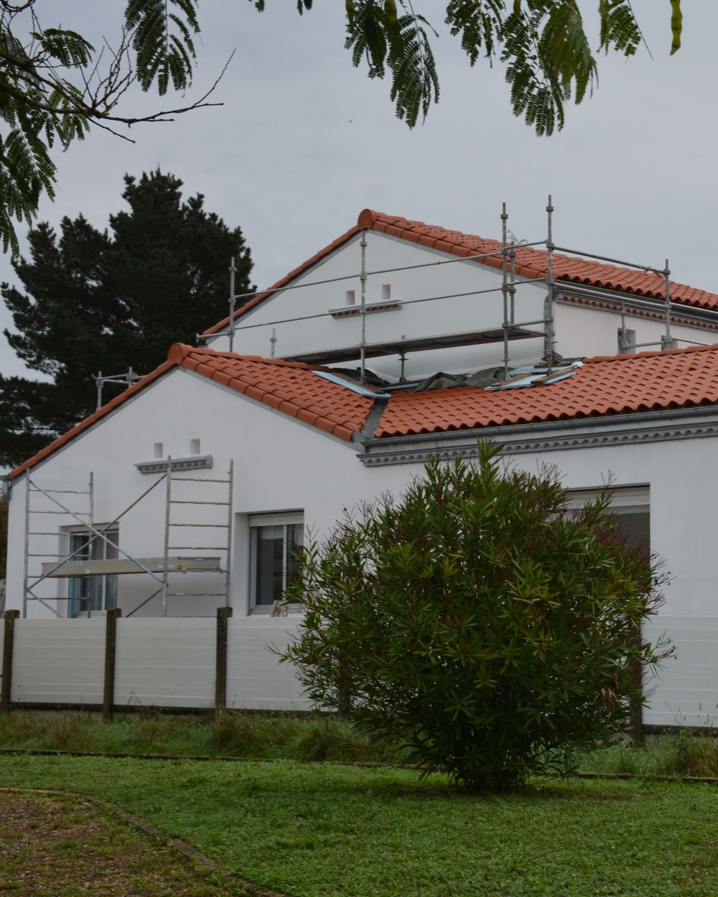 Bâtiment blanc au toit de tuiles rouges, échafaudages et un petit buisson devant.