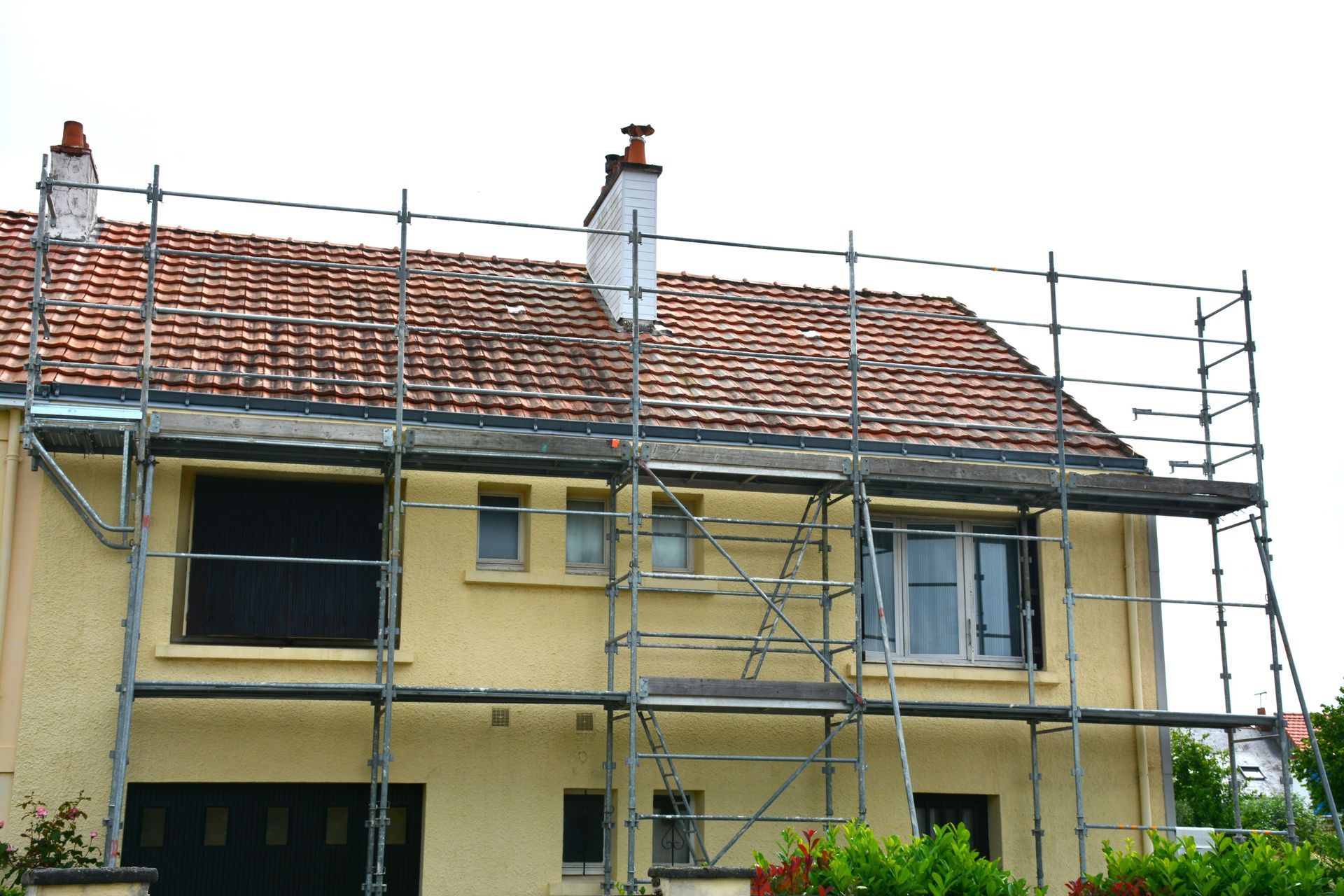 Des échafaudages sont installés autour d'une maison beige à deux étages avec un toit de tuiles rouges.