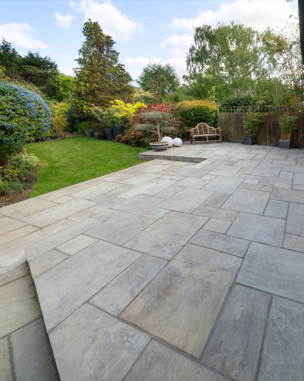 Terrasse en pierre donnant sur un jardin avec pelouse, arbres et banc.