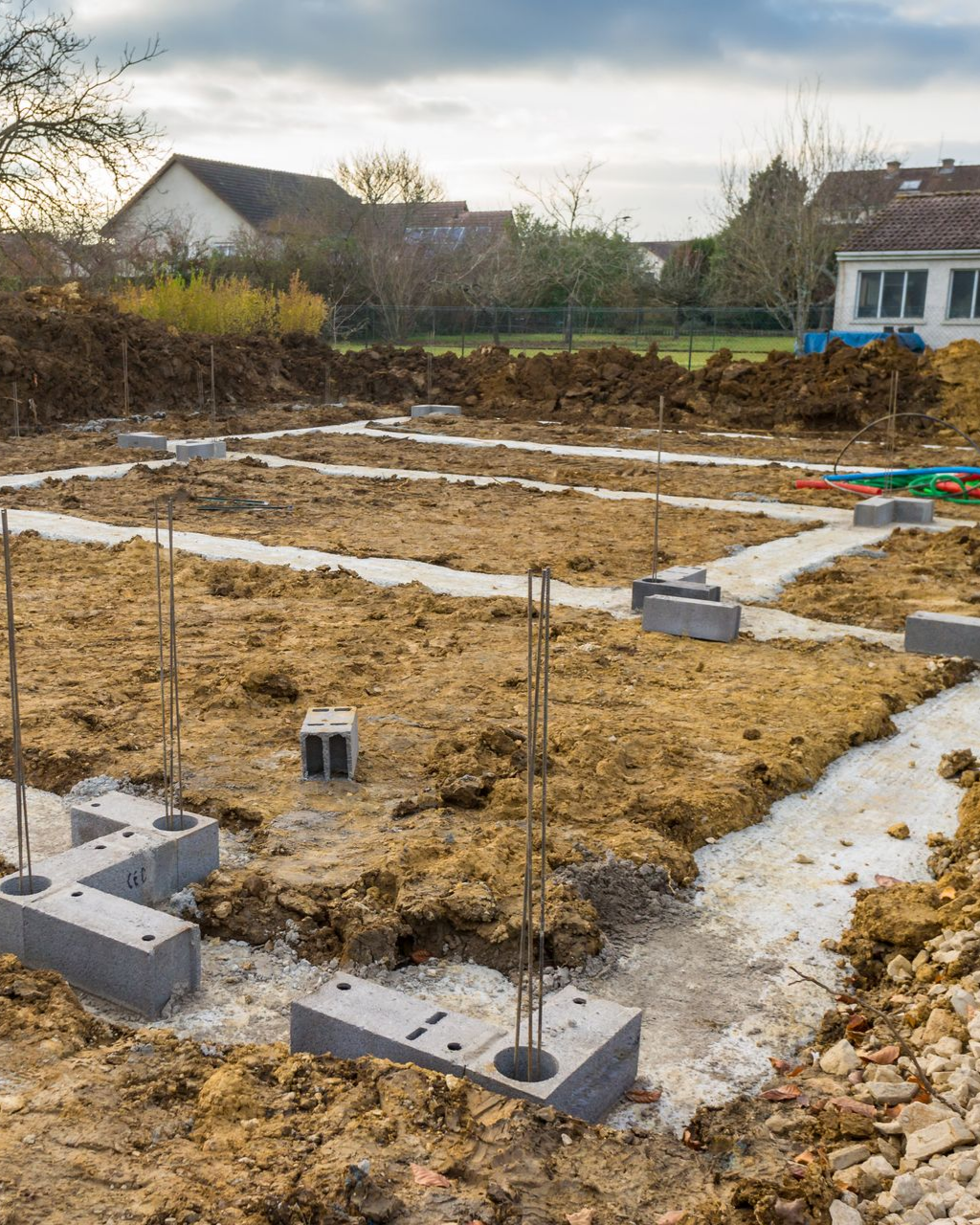 Chantier : blocs de fondation en béton et barres d'armature posés sur la terre, avec des maisons en arrière-plan.