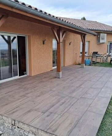 Terrasse couverte avec poutres en bois et sol carrelé. Bâtiment avec porte-fenêtre coulissante et barbecue extérieur.