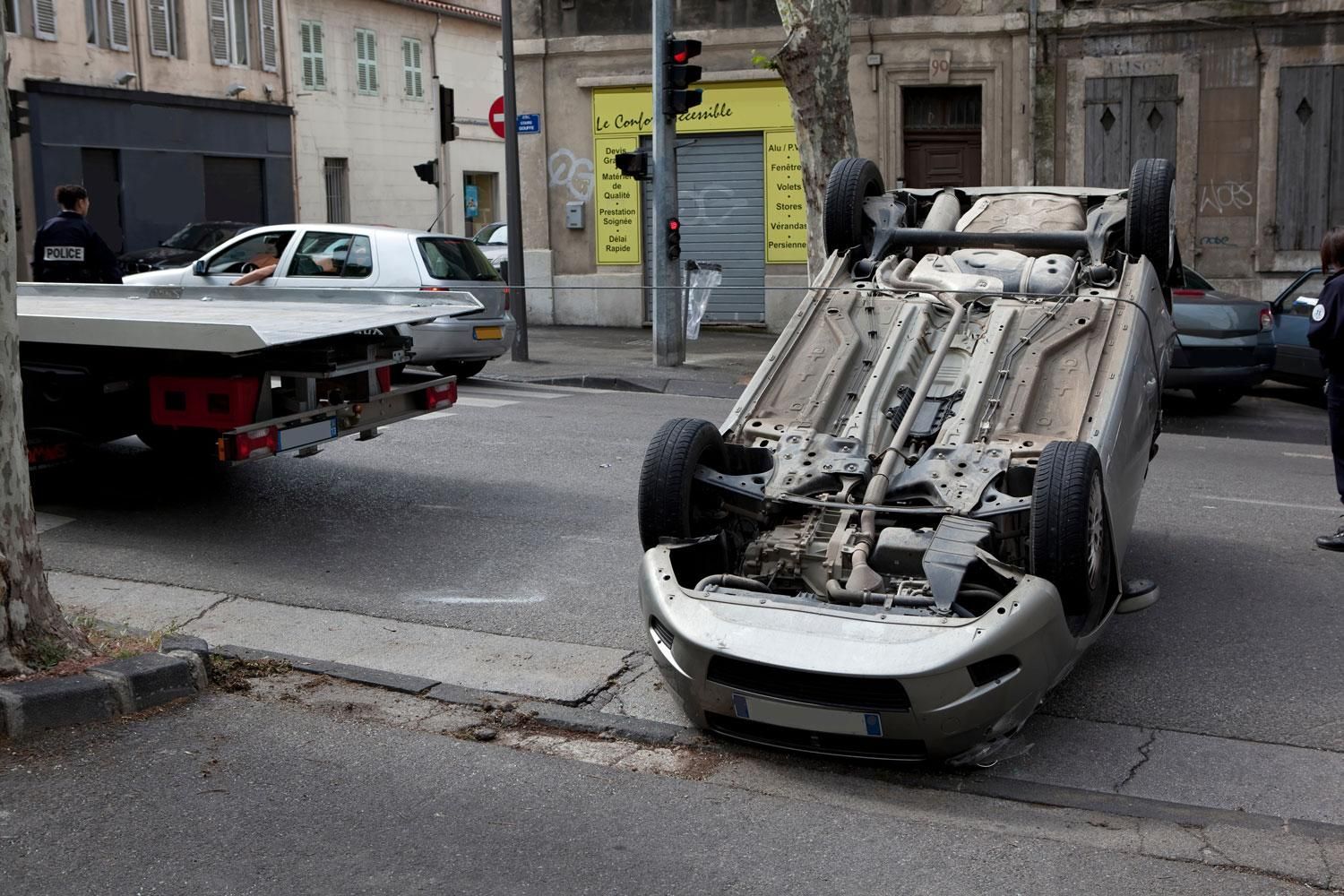 Voiture blanche renversée dans une rue en ville, à côté d’une dépanneuse, avec des débris à proximité.