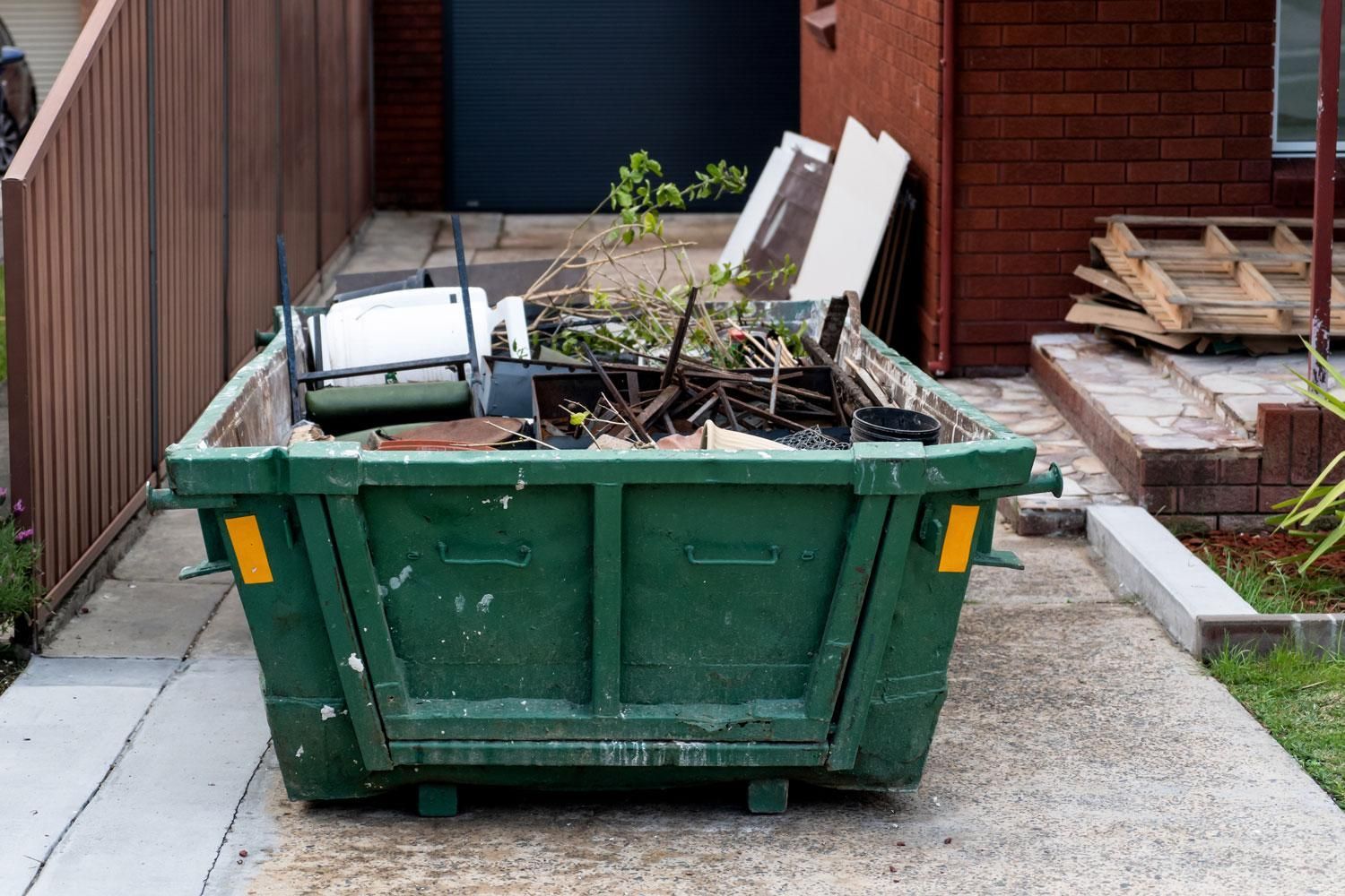 Benne de chantier verte remplie de débris et de déchets verts, à côté d’un bâtiment et d’un escalier en bois