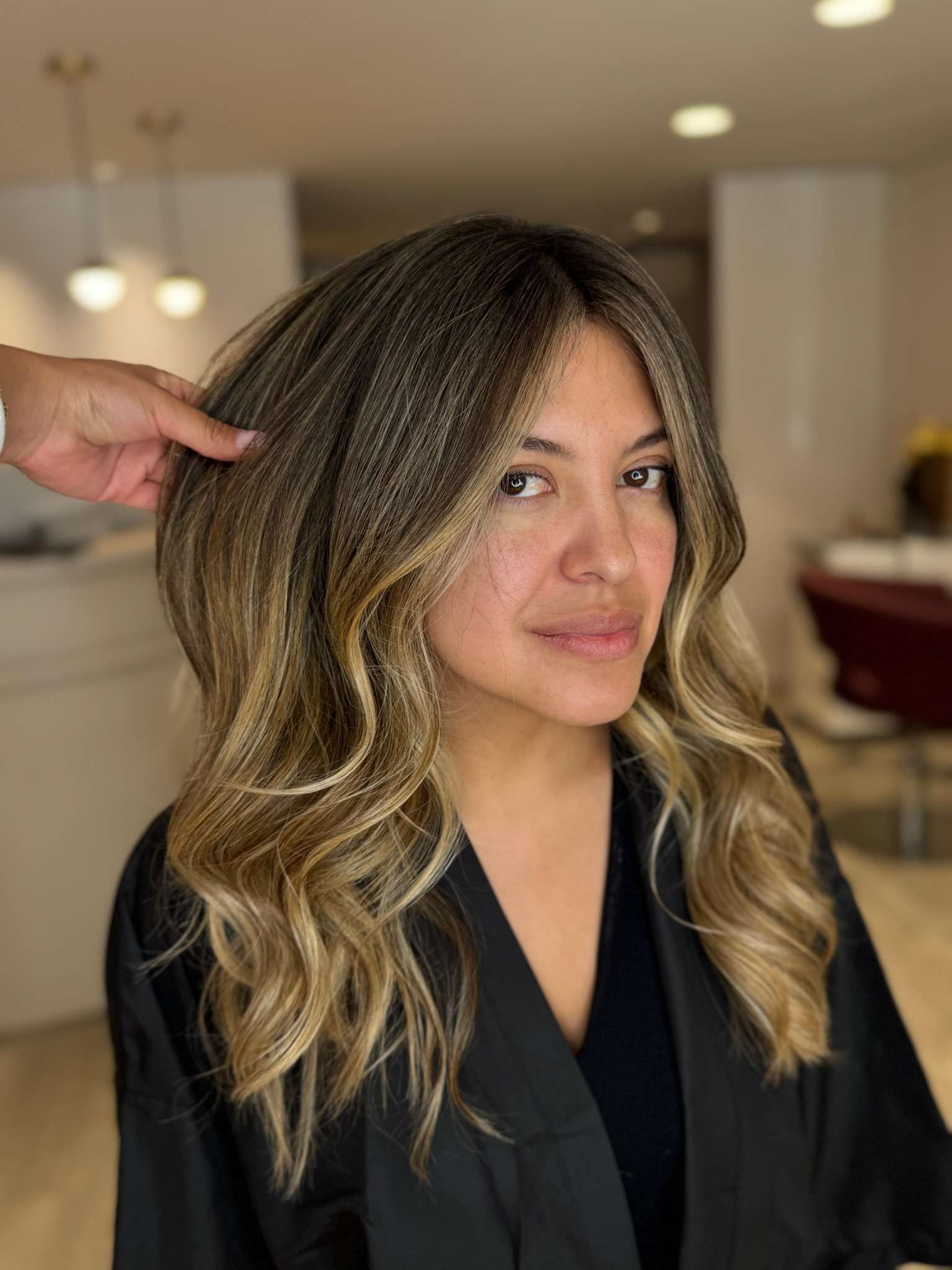 Mujer con cabello ondulado con mechas, vestida con una capa negra de peluquería, en una peluquería.