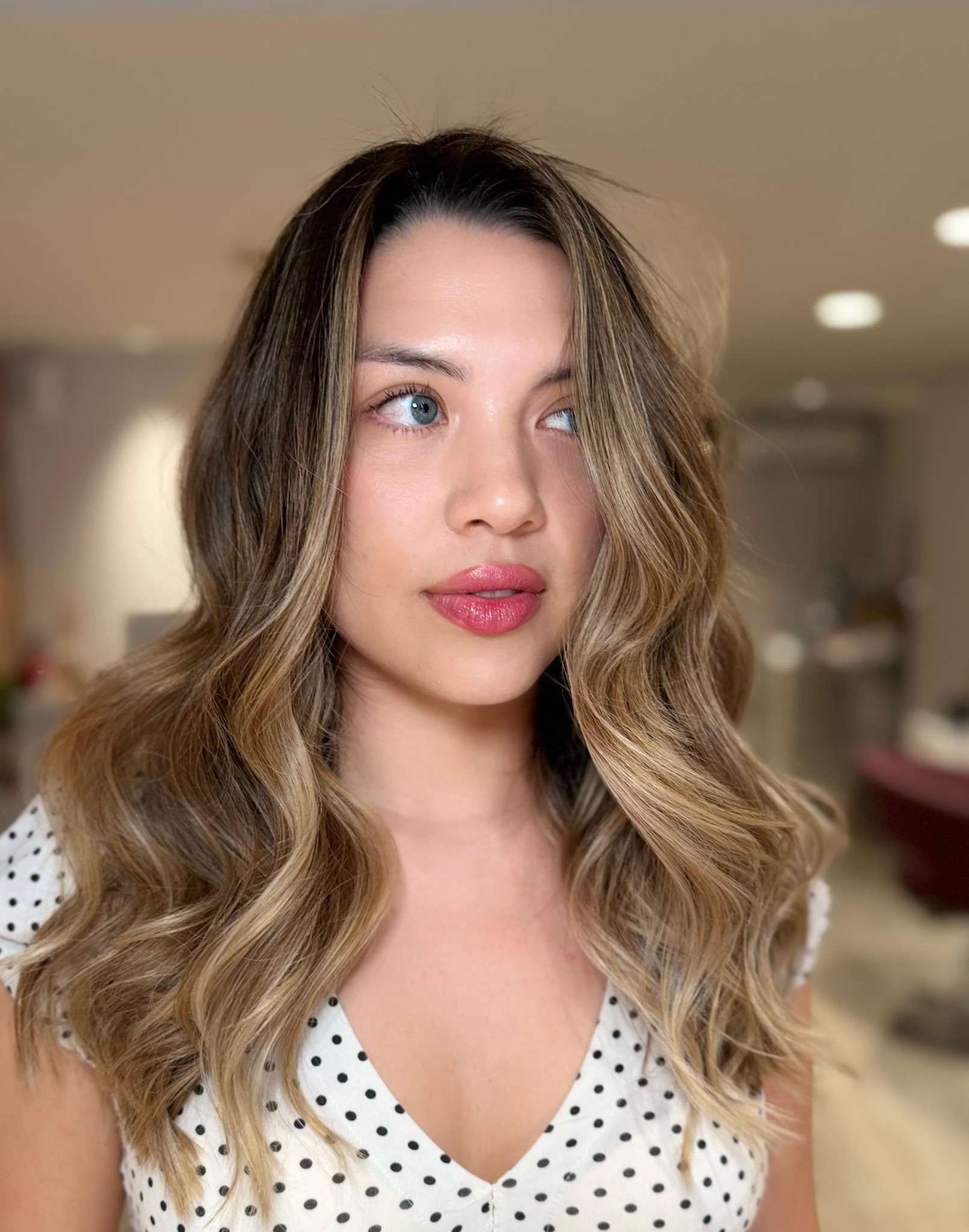 Mujer con cabello ondulado y mechas, vestida con una blusa blanca de lunares, mirando de reojo.