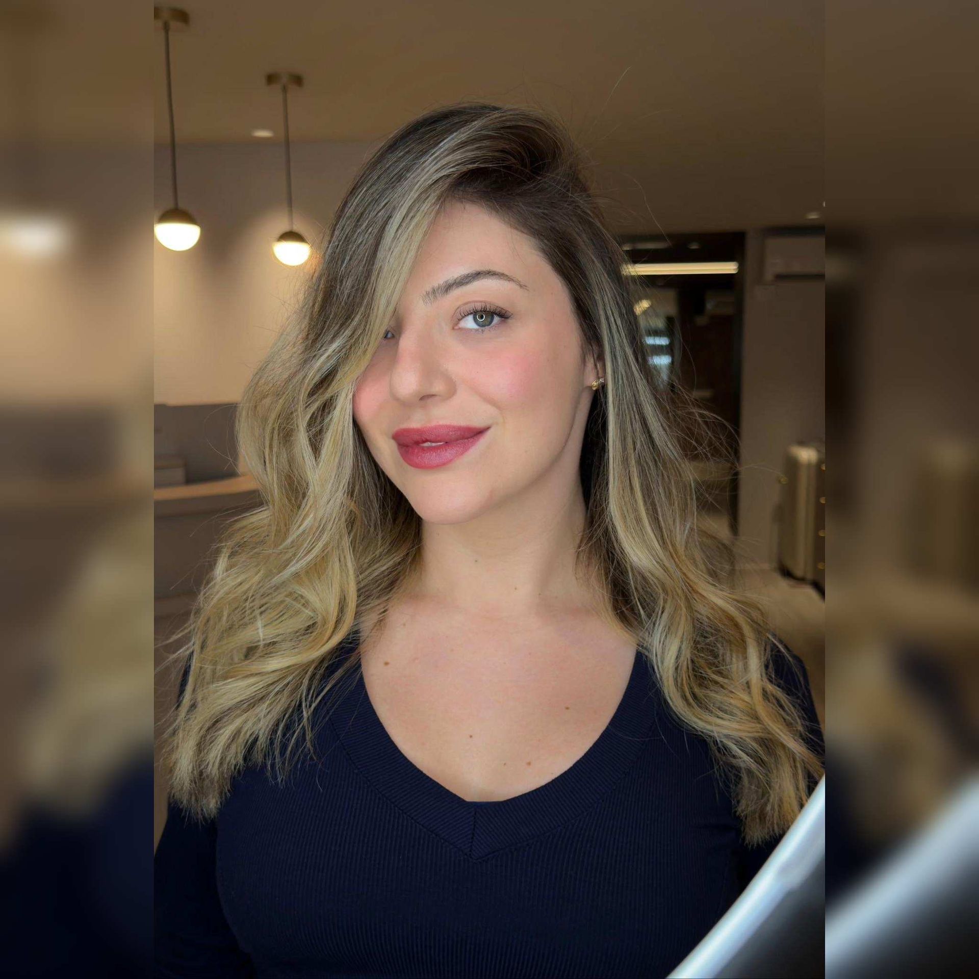 Mujer con cabello rubio largo y lápiz labial rosa, sonriendo en un ambiente interior con iluminación cálida.