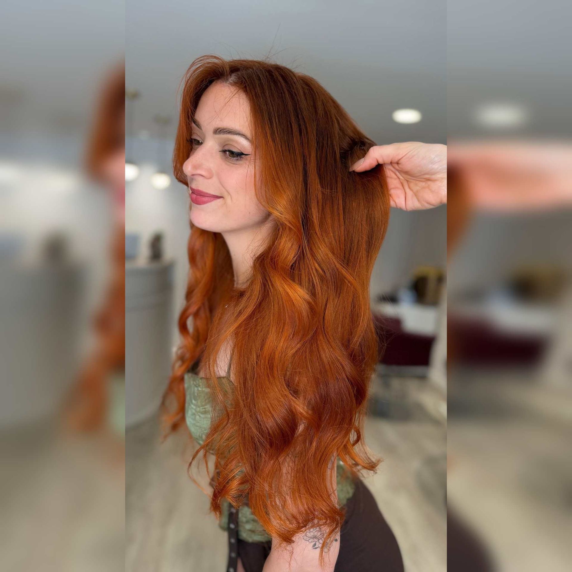 Mujer con cabello largo, ondulado y rojo en una peluquería, con una mano levantándose el cabello para mostrar su textura.