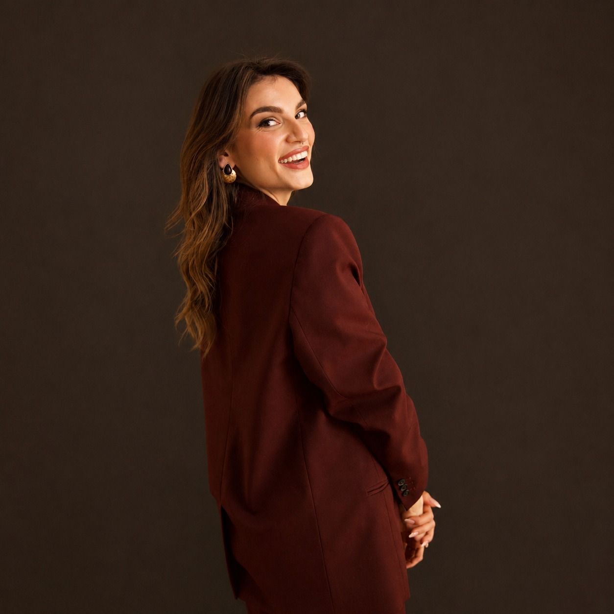 Mujer con traje burdeos sonriendo por encima del hombro contra un fondo de estudio oscuro