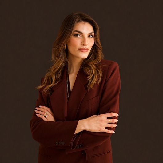 Retrato de una mujer con un blazer color burdeos y los brazos cruzados sobre un fondo oscuro de estudio.