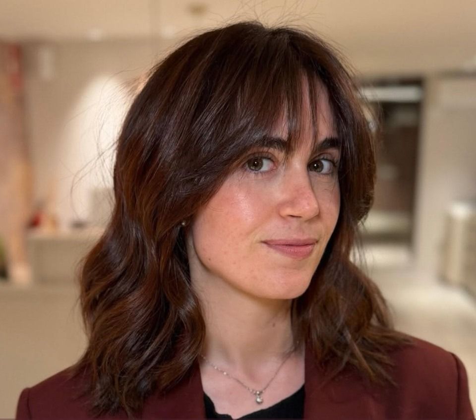 Retrato de una persona con cabello castaño ondulado, vestida con un blazer granate, de pie en un interior con un fondo suave.