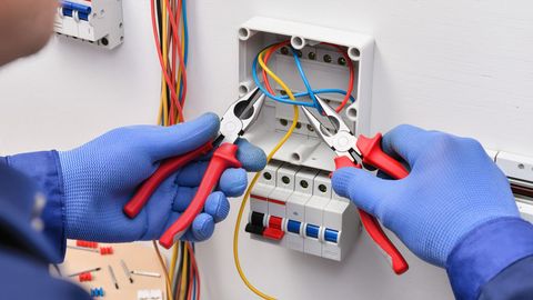 Eine Person mit  arbeitet mit einer Zange an elektrischen Drähten in einer offenen, an der Wand montierten Anschlussdose.