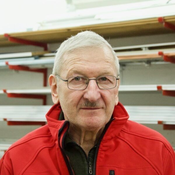 Eine Person mit Brille und roter Jacke steht vor Regalen.