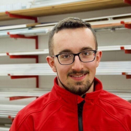 Eine Person mit Brille und Bart, die eine rote Reissverschlussjacke trägt, steht in einer Lagerhalle mit Metallregalen.