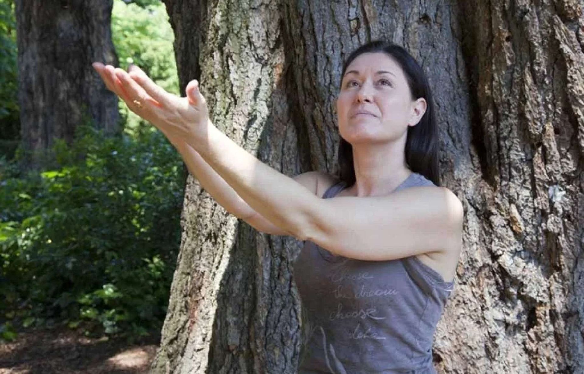 Femme les bras levés devant un arbre, regardant vers le ciel. En extérieur, temps ensoleillé.