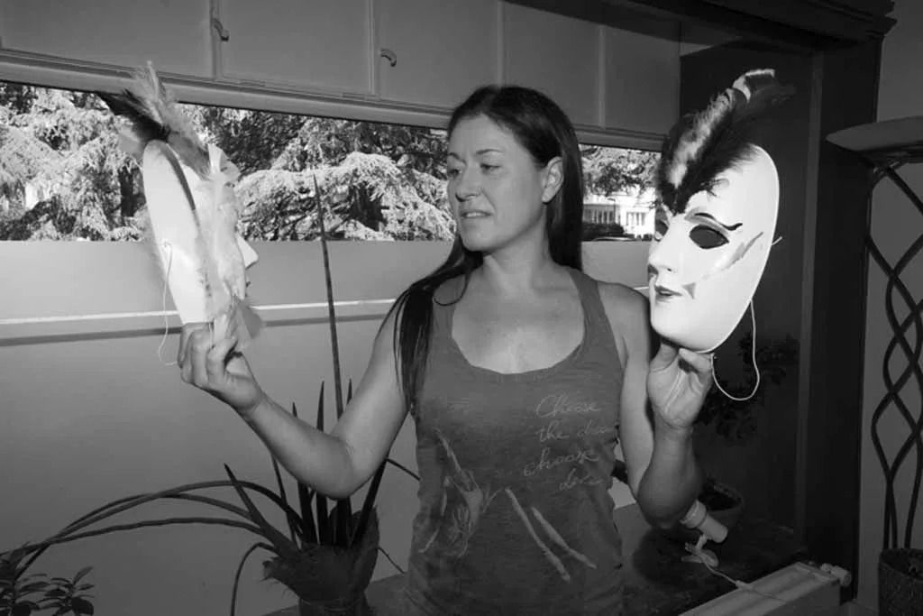 Femme tenant deux masques décoratifs, l'un blanc et l'autre avec une perruque noire, près d'une fenêtre.