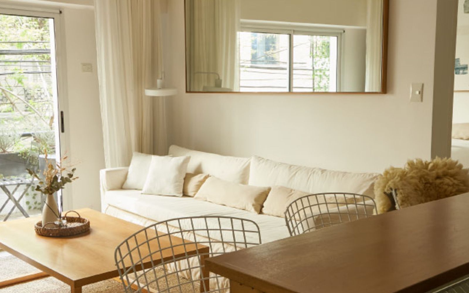 Luminosa sala de estar con sofá blanco, mesa de madera y sillas de alambre. Espejo en la pared y puerta al patio.