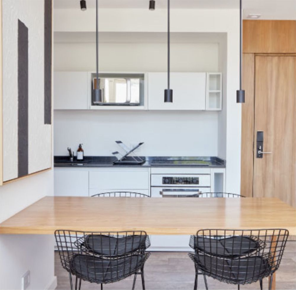 Cocina y comedor con modernas luces colgantes negras, mesa de madera clara y sillas de alambre.