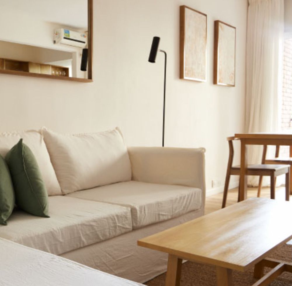 Sala de estar con sofá crema, almohadas verdes, mesa de madera, espejo y dos obras de arte enmarcadas.