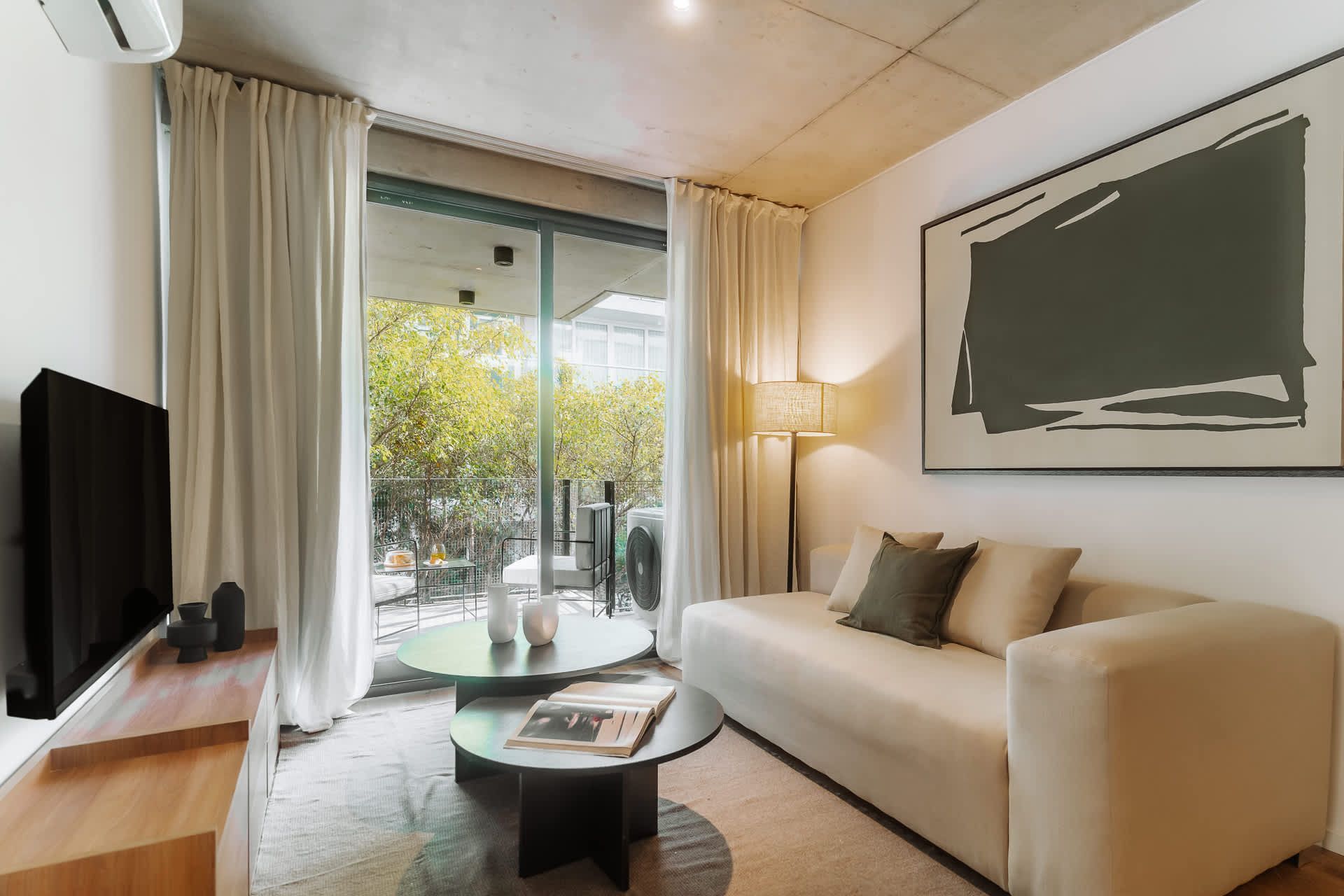 Un salón de tonos neutros con un televisor montado en la pared, dos estanterías de madera, dos mesas pequeñas y sofás de color crema.