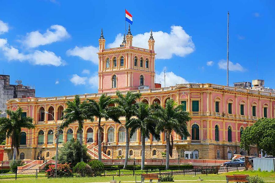 Palacio Presidencial en Asunción, Paraguay, edificio rosa con bandera en la parte superior, palmeras al frente, cielo azul.