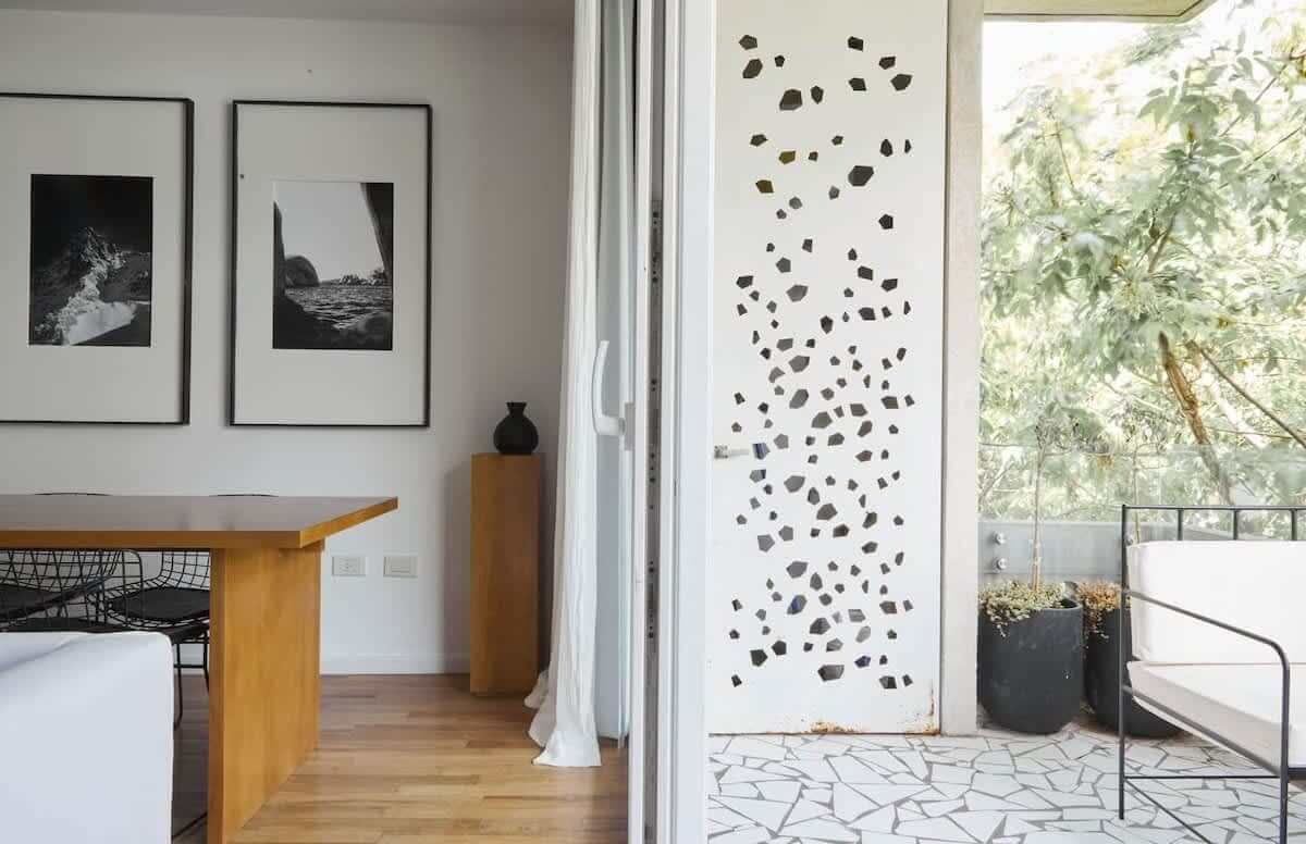 Sala de estar con sofá beige, obras de arte, mesas de café y una vista de la vegetación a través de una puerta de vidrio.